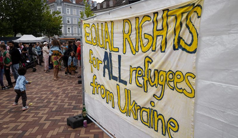 Banner saying EQUAL RIGHTS FOR ALL REFUGEES FROM UKRAINE and people in the background