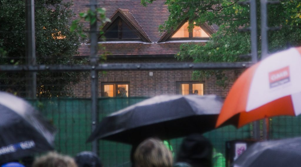 The Manston camp in the background, with illuminated windows, in which a family with a baby can be seen in one. Protesters and umbrellas in foreground, a guarded fence in the middle ground.