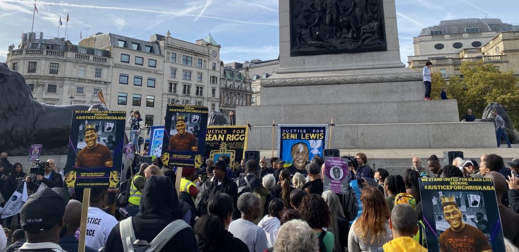 Trafalgar Square, Nelson's column in the background and buildings, and a large crowd of protesters holding placards and banners commemorating dead victims of police and state violence.
