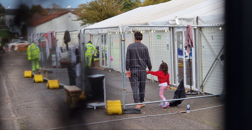 A man holding hands with his daughter inside Manston camp, glimpsed through the gates. There are security officers in the background and two white marquees.