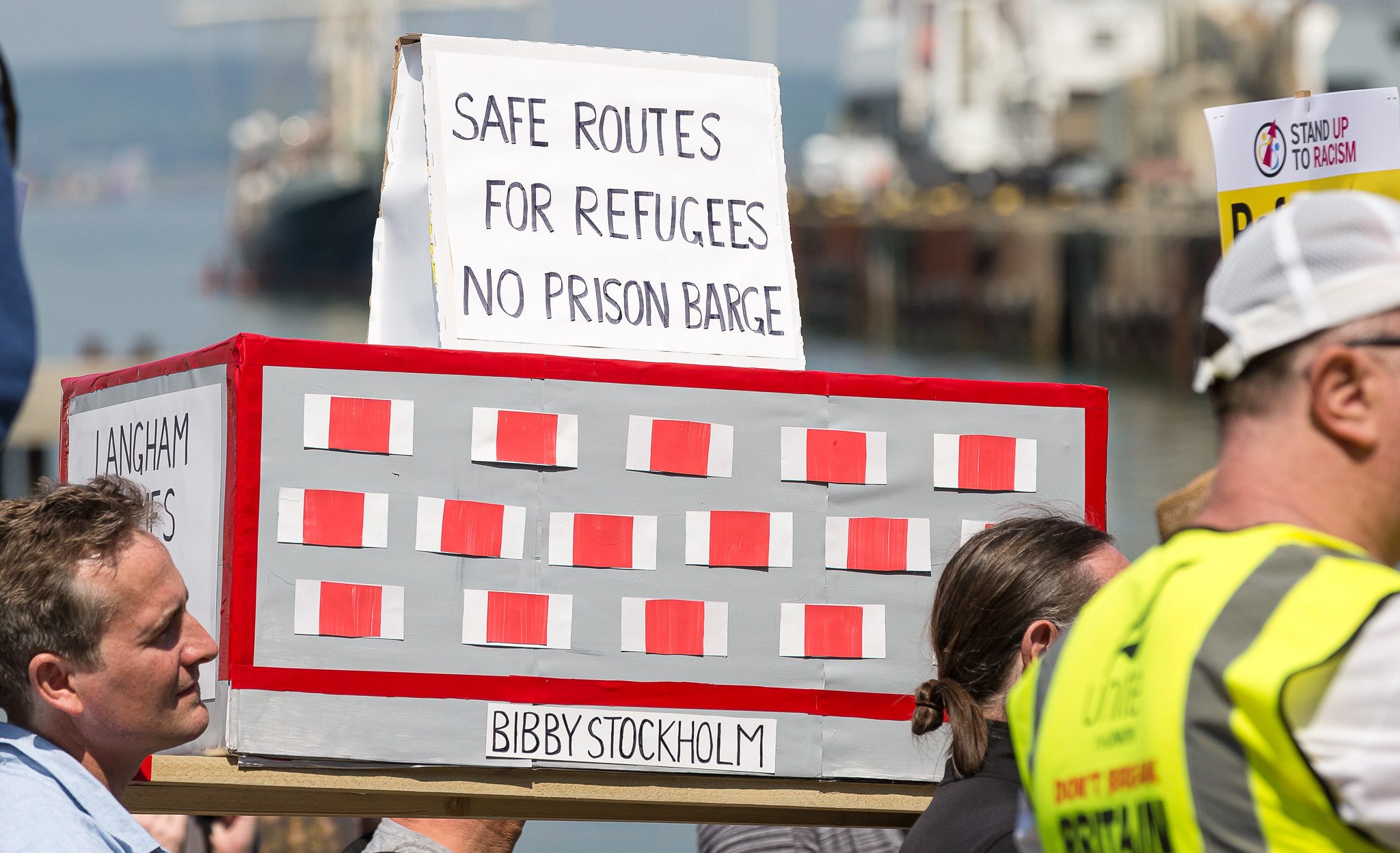 Placard saying safe routes for refugees, no prison barge, on top of a protest mock barge called Bibby Stockholm, at protest in Dorset against refugee barge and in solidarity with refugees.